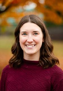 Ellie Willemin headshot; wearing a maroon sweater with a colorful fall backdrop, and smiling
